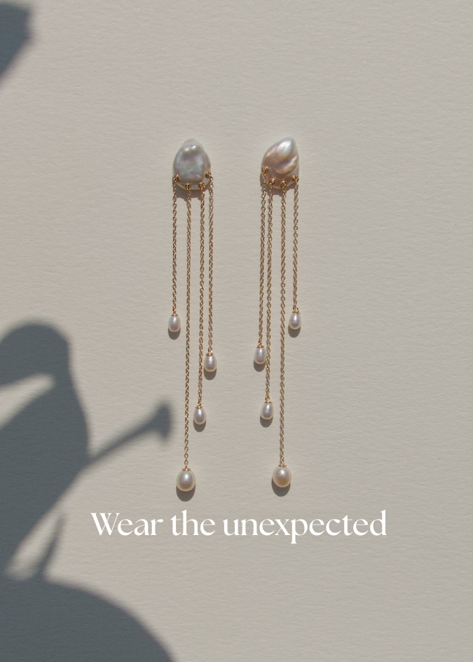 Gold earrings with pearl drops displayed on a neutral background and worn by a person.