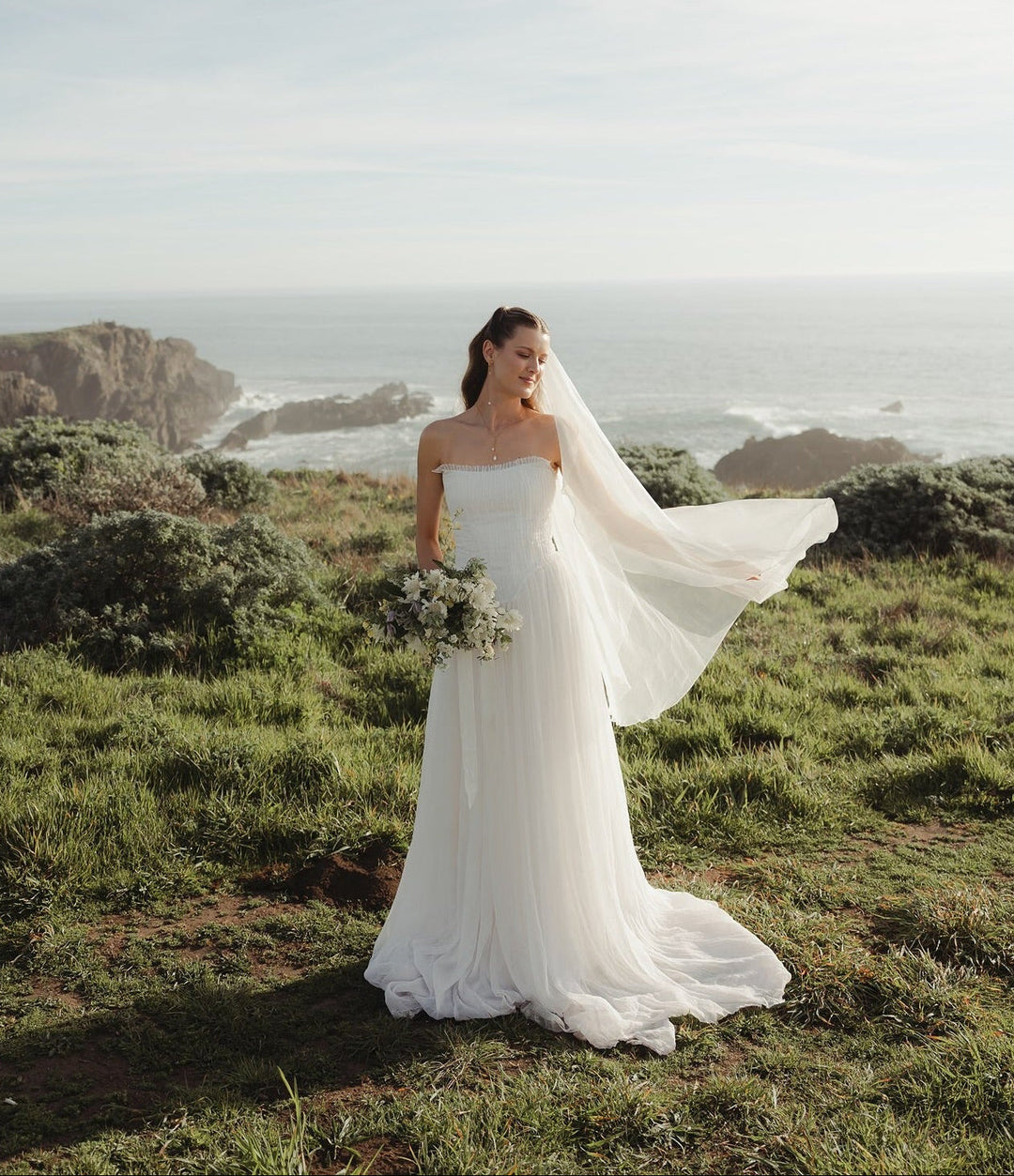 Woman in a white wedding dress standing on a grassy hill with ocean view
