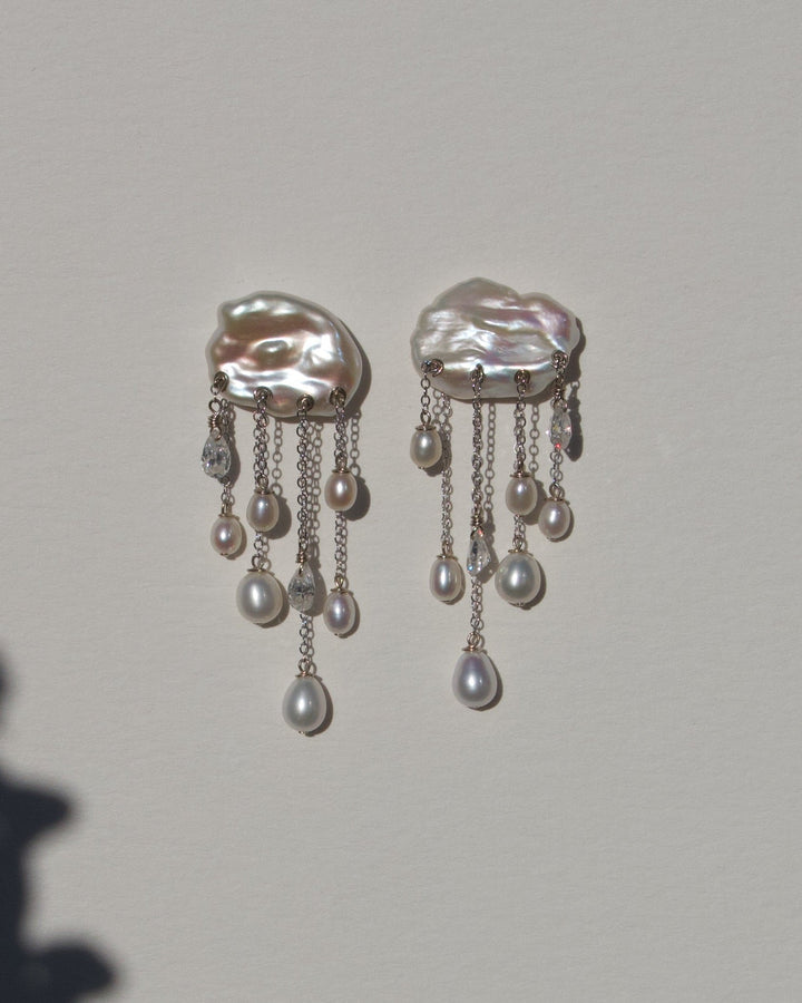 Halo white keshi and teardrop pearl earrings with diamonds and 14k white gold chains, displayed against a wall with floral shadows.