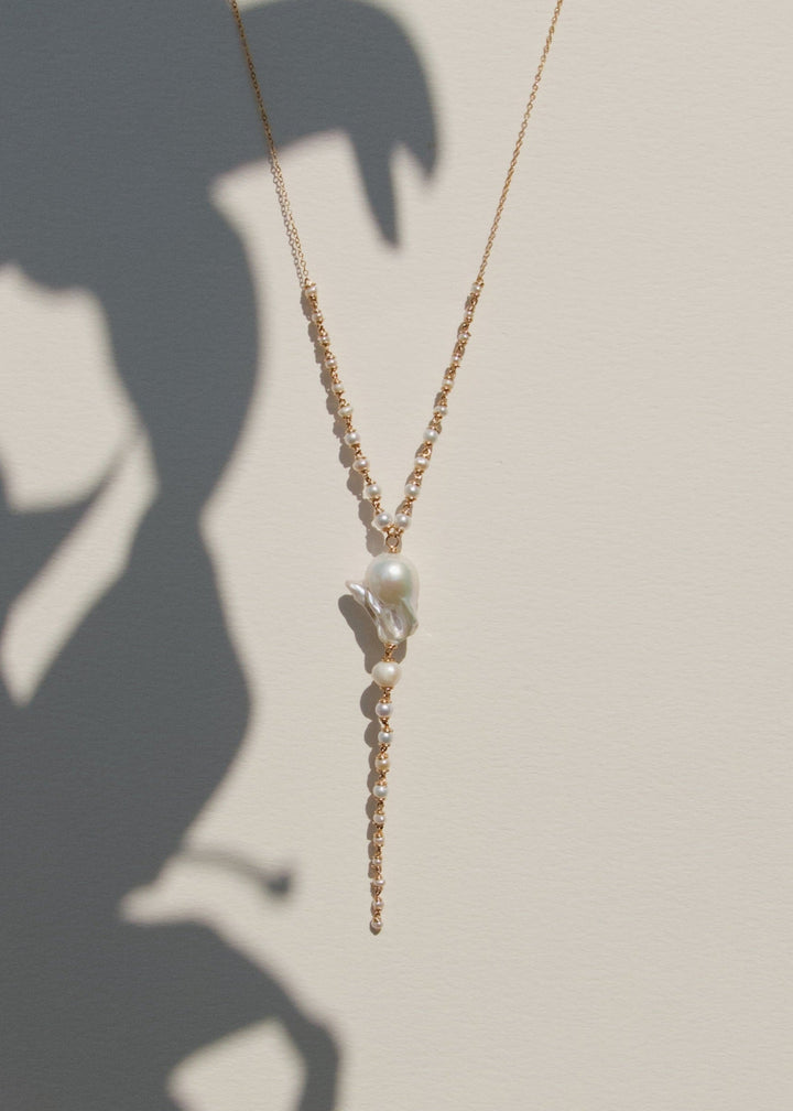 Mira - This lariat necklace is all about balance between structure and softness, simplicity and detail. A luminous baroque pearl anchors the center, framed by three strands of seed pearls that taper gently in size. They meet a delicate chain that adds jus