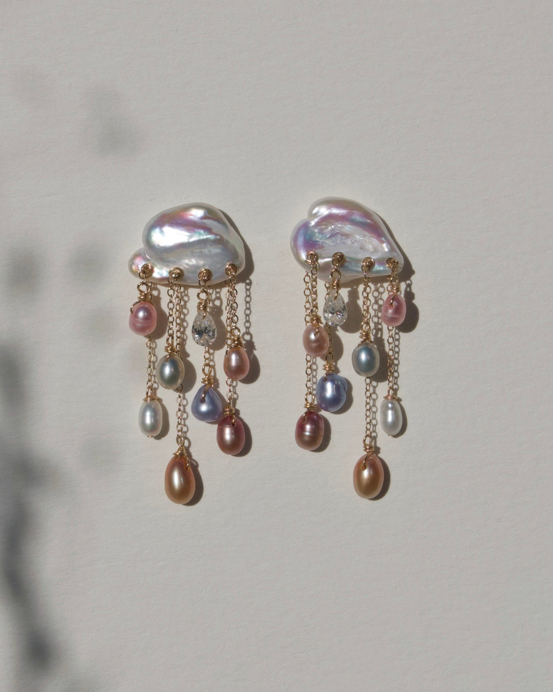 Close-up of Stardust earrings with iridescent keshi pearls, pink, peach and lavender pearl drops and diamonds on gold chains beside plant shadows.